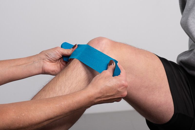 Person appliziert blaues Kinesiotape auf ein Knie.