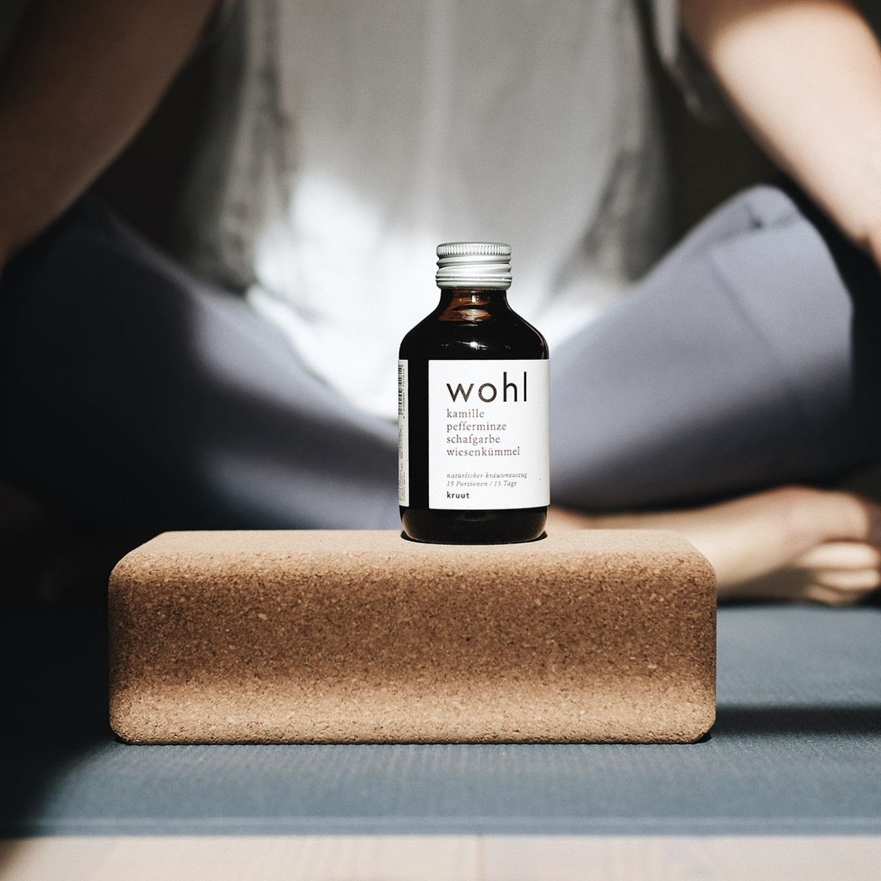 Braune Glasflasche mit silbernem Deckel auf einem Yogablock. Aufschrift: wohl, Kamille, Pfefferminze, Schafgarbe, Wiesenkümmel, kruut. Person im Hintergrund.