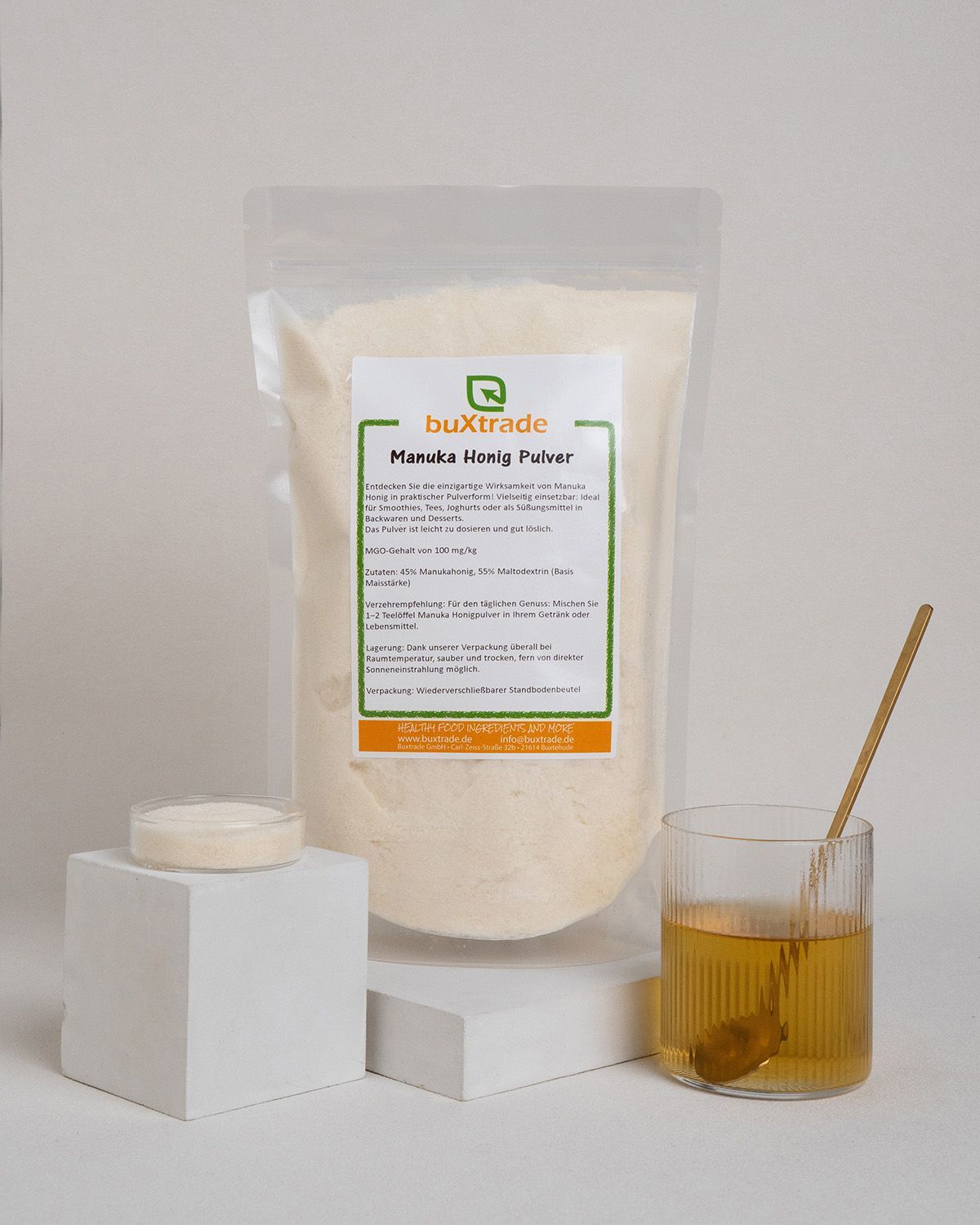 Beutel mit Pulver, Glas mit Flüssigkeit, Löffel, Podest, Etikett 'buXtrade Manuka Honig Pulver'.