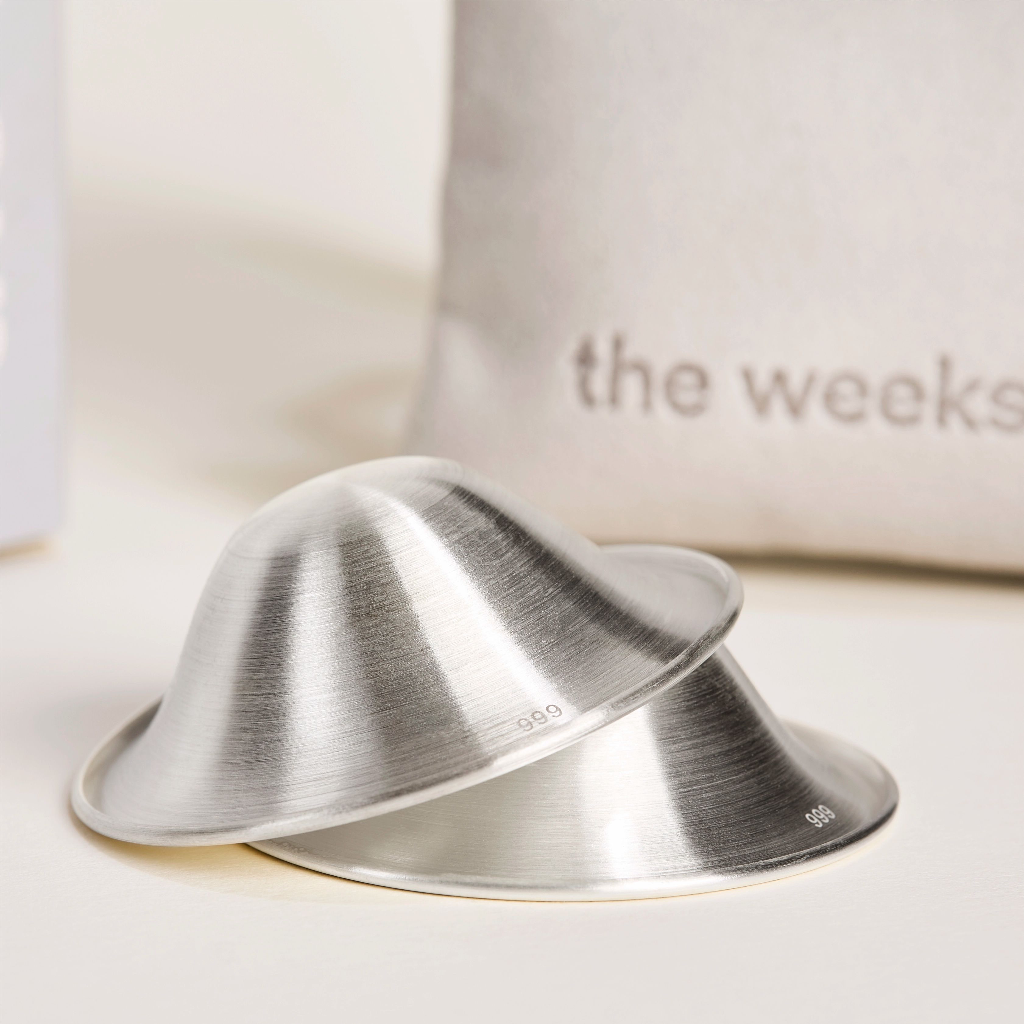 Zwei silberne Stillhütchen, leicht überlappend. Im Hintergrund ein Beutel mit Logo.