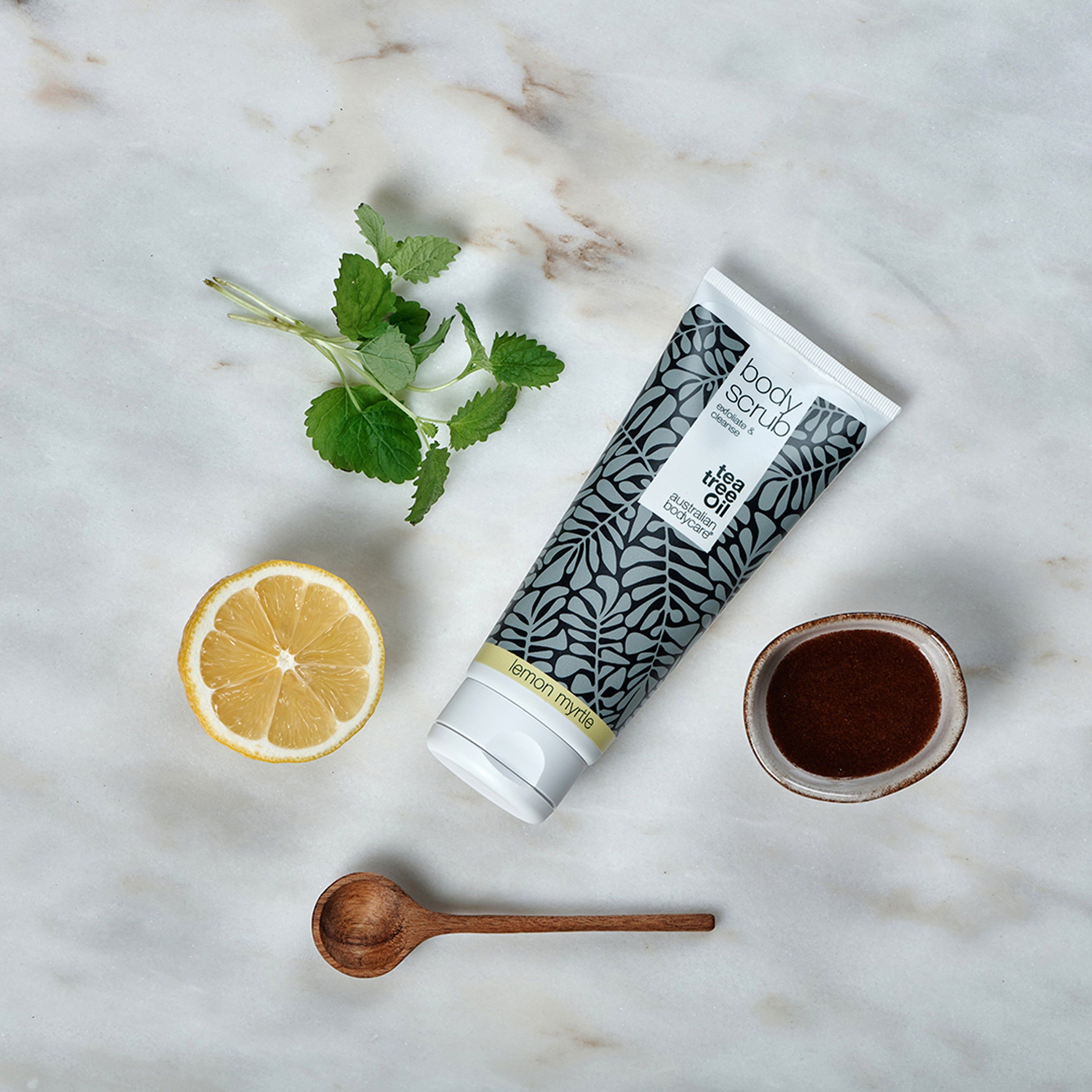 Körperpeeling-Tube, Zitrone, Minze und Holzlöffel auf Marmor. Aufschrift: Body Scrub, Australian Bodycare.