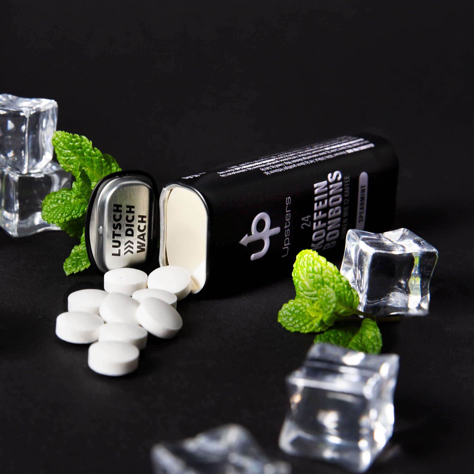 Geöffnete schwarze Dose mit weißen Tabletten. Aufschrift: Upsters, 24 Koffein Bonbons, Spearmint. Daneben Minze und Eiswürfel.