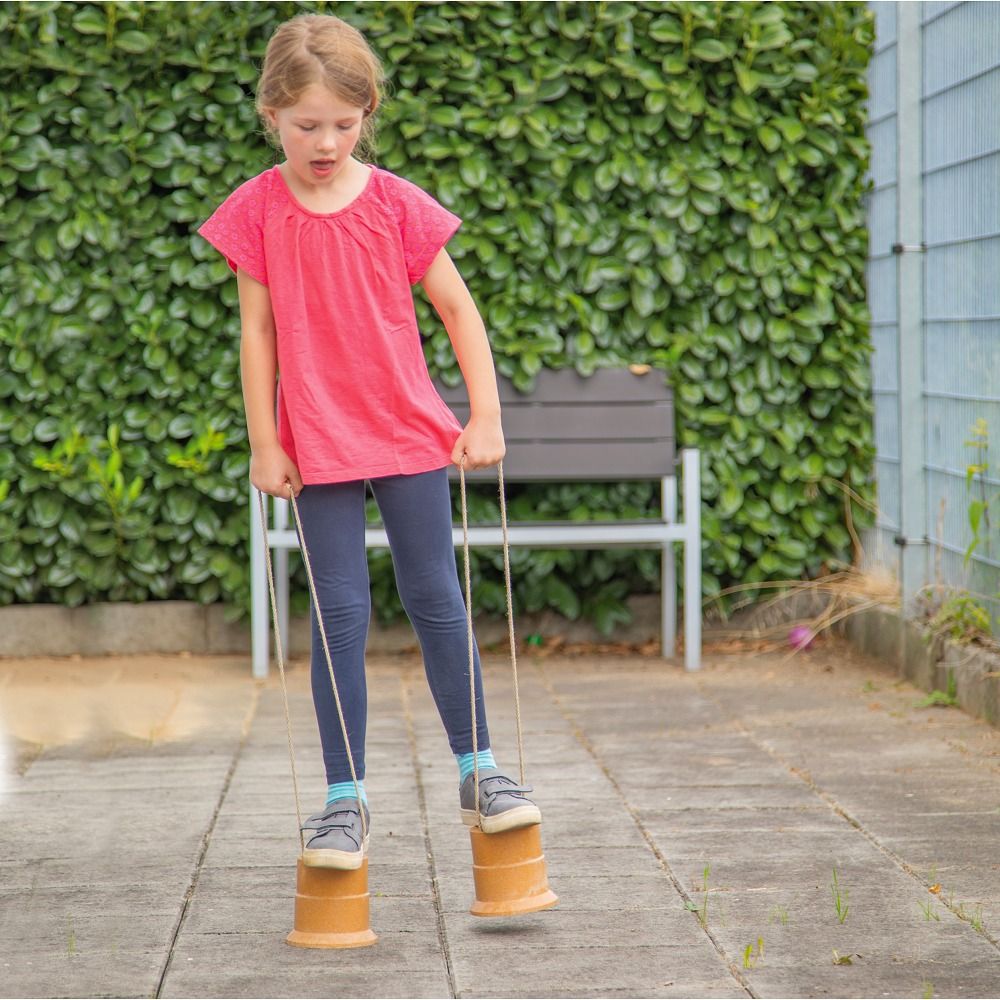 Kind auf Laufdosen. Beige Dosen mit Schnüren. Kind trägt rotes Shirt und blaue Hose. Hintergrund: Hecke und Bank.