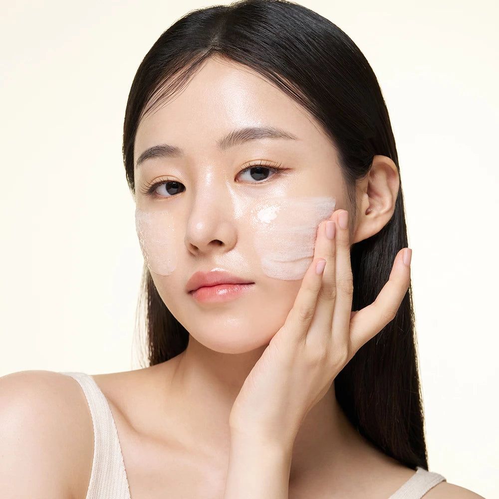 Eine Frau trägt ein weißes Top. Sie trägt eine transparente Substanz auf ihrem Gesicht auf.