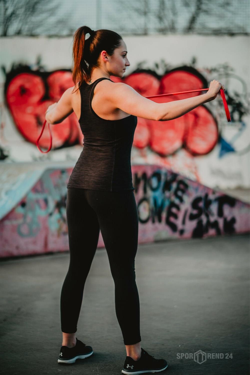 Frau trainiert mit rotem Fitnessband im Freien. Sie steht seitlich und hält das Band.