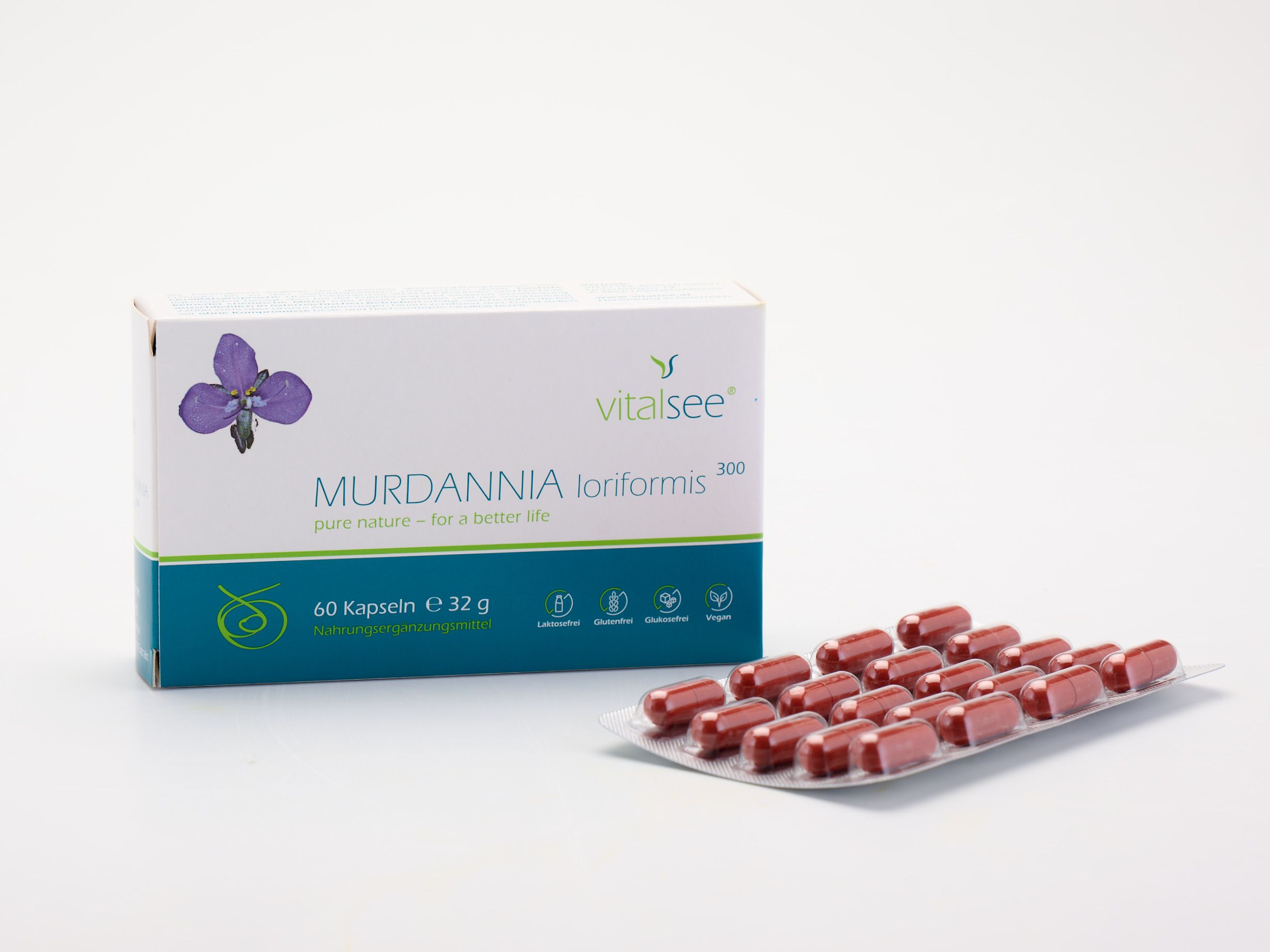 Weiße Schachtel mit lila Blüte und Text. Daneben liegt eine Blisterpackung mit roten Kapseln. Aufschrift: Vitalsee, MURDANNIA loriformis 300 Extrakt.