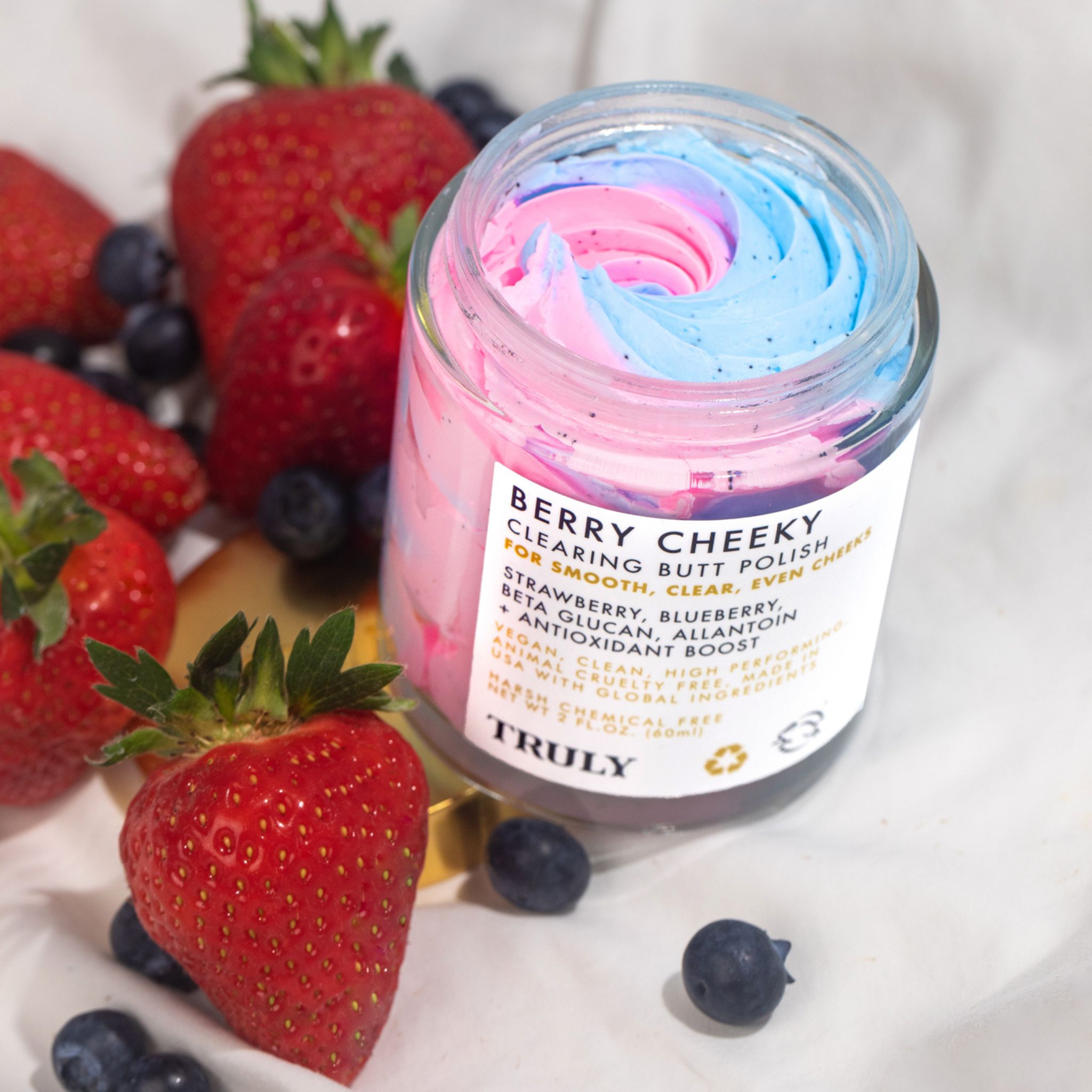 Glasbehälter mit rosa und blauem Produkt, umgeben von Erdbeeren und Blaubeeren. Produktetikett sichtbar.