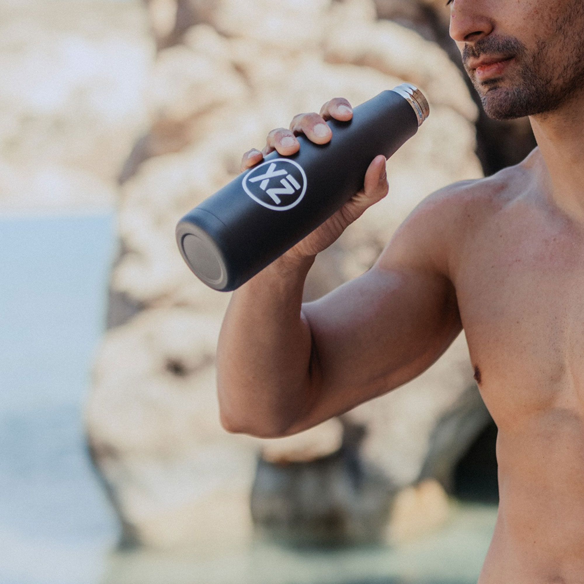 Mann hält schwarze Isolier-Trinkflasche. Logo sichtbar. Hintergrund: Felsen und Meer.
