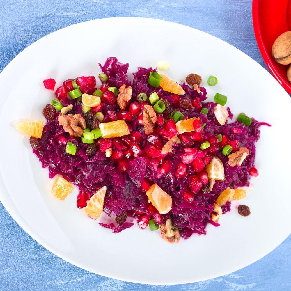 Teller mit Rotkohlsalat. Zutaten: Granatapfelkerne, Walnüsse, Rosinen, Mandarinenstücke und grüne Zwiebeln. Blauer Hintergrund.
