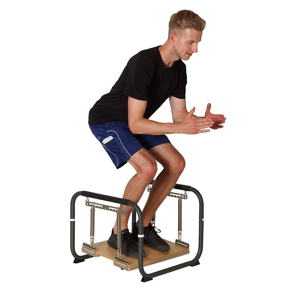 Mann steht auf Trainingsgerät. Schwarzer Rahmen, Holzplatte, Federn und Griffe. Mann in Sportkleidung. Weißer Hintergrund.