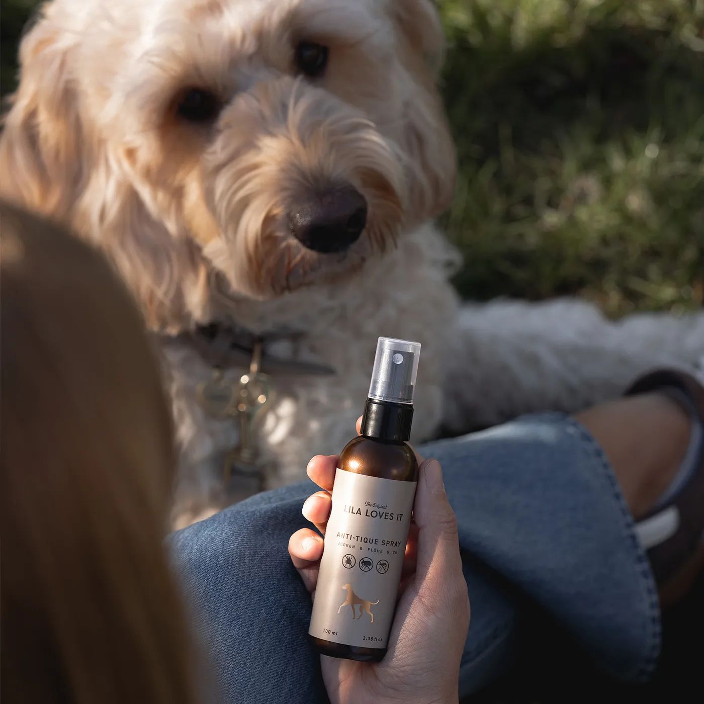 Person hält eine braune Sprühflasche mit schwarzem Sprühkopf. Aufschrift: LILA LOVES IT, Anti-Zecken Spray. Hund im Hintergrund.