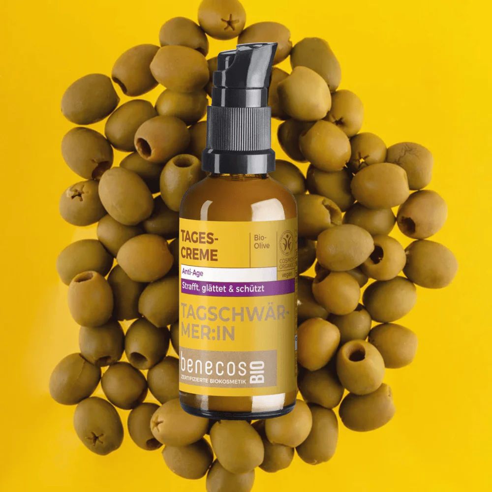 benecosBIO Tagescreme BIO-Olive Flakon umgeben von Oliven auf gelbem Hintergrund.