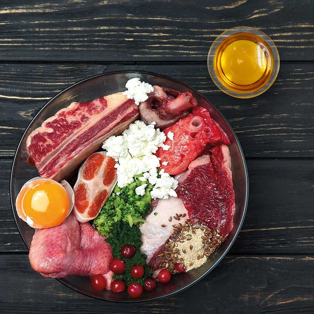 Glasplatte mit verschiedenen Lebensmitteln: Fleisch, Eigelb, Gemüse, Öl. Auf dunklem Holzuntergrund.