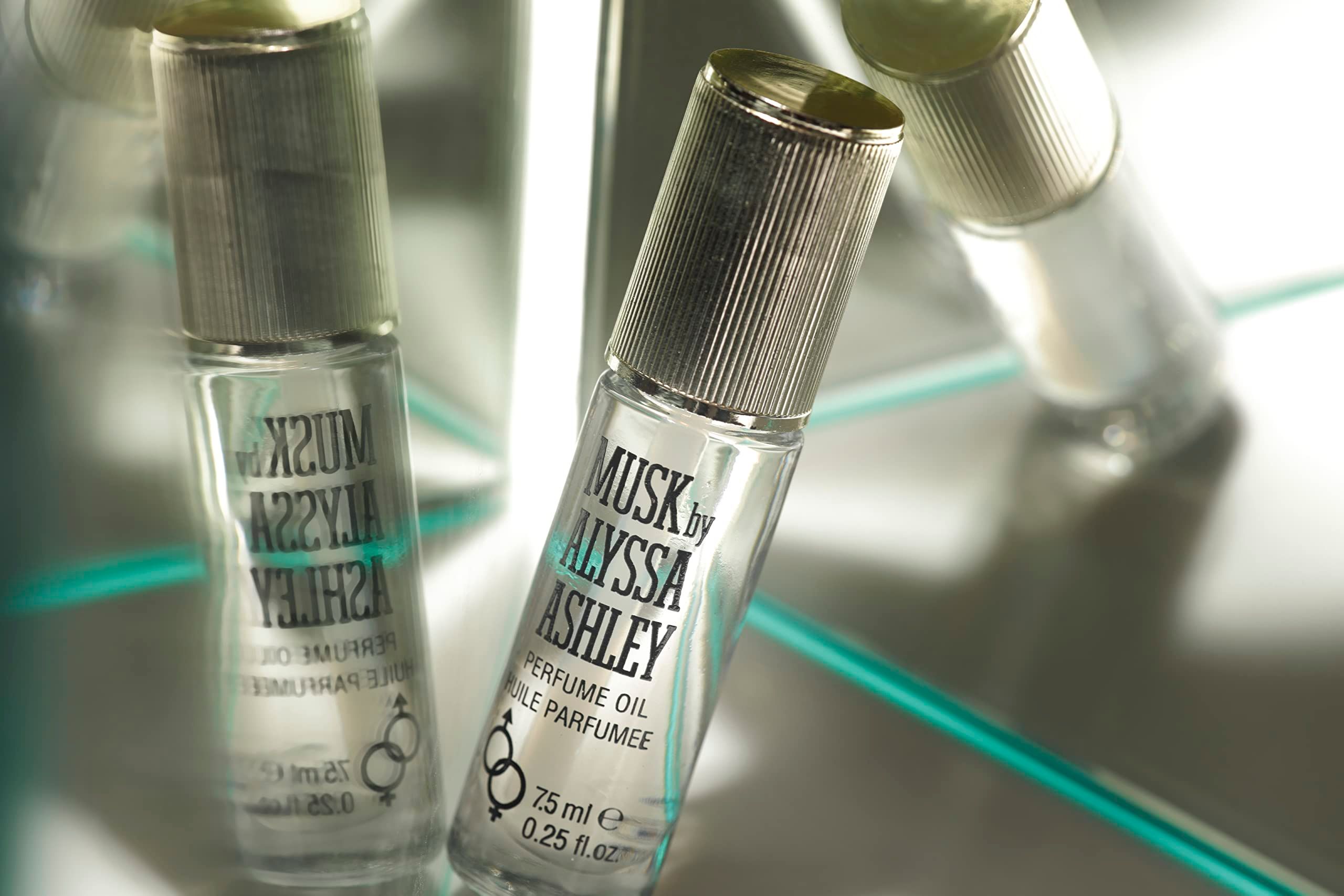 Zwei Alyssa Ashley Musk Parfumöl Flakons auf spiegelnder Oberfläche, mit goldenen Deckeln.