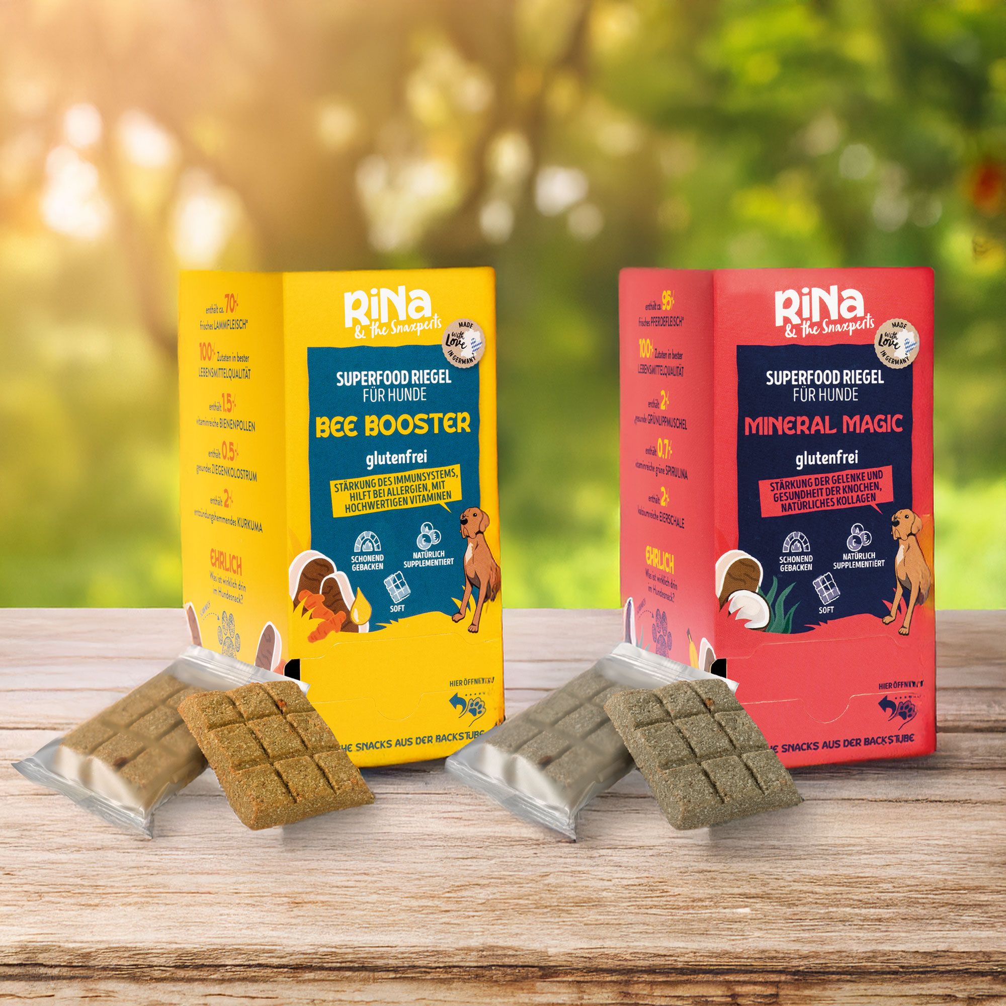 Zwei Verpackungen mit Hundesnacks. Aufschrift: RiNa, Superfood Riegel, BEE BOOSTER und Mineral Magic, glutenfrei. Snacks daneben.