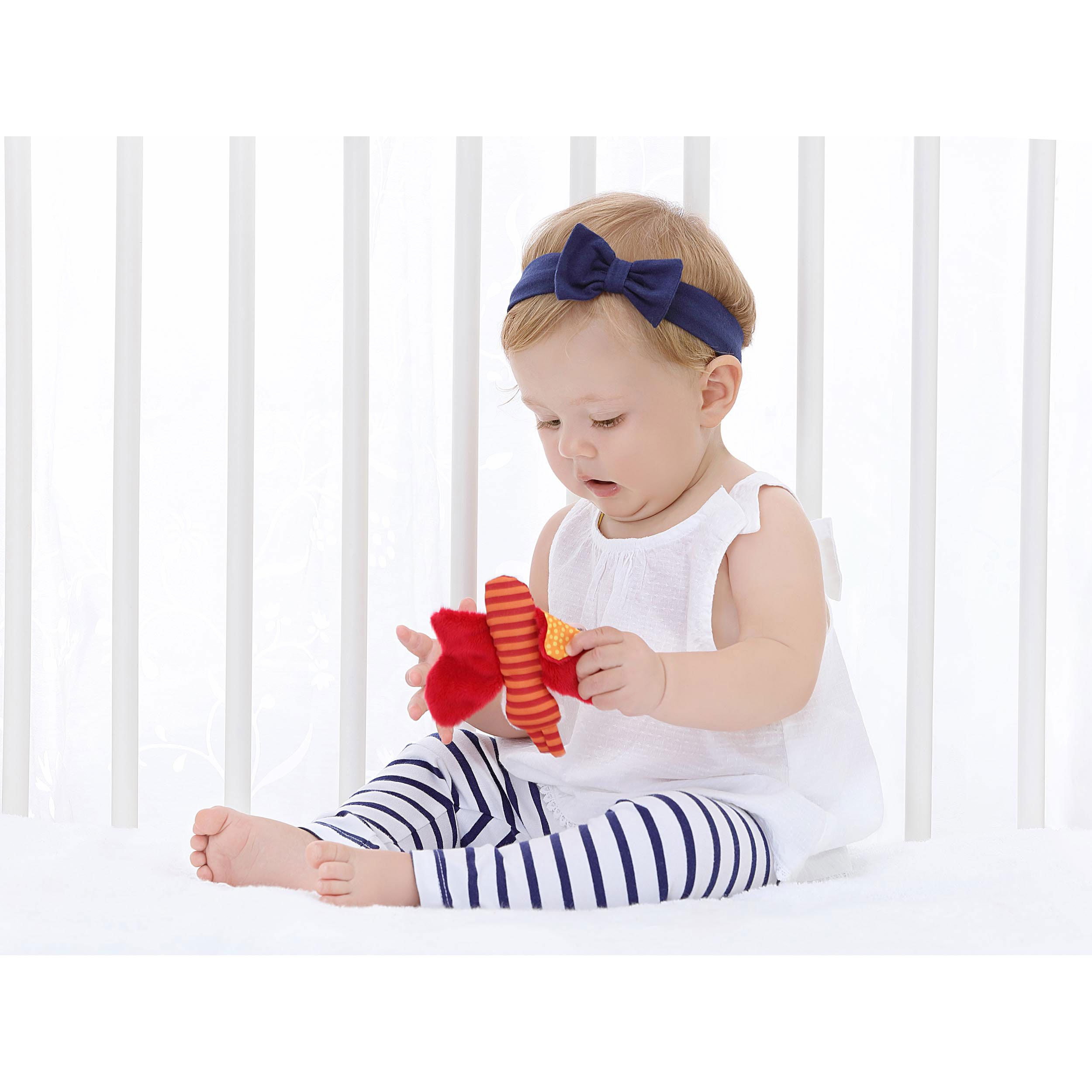 Baby hält ein rotes und orangefarbenes Schmetterlingsspielzeug. Das Kind trägt ein blaues Stirnband und gestreifte Hose.
