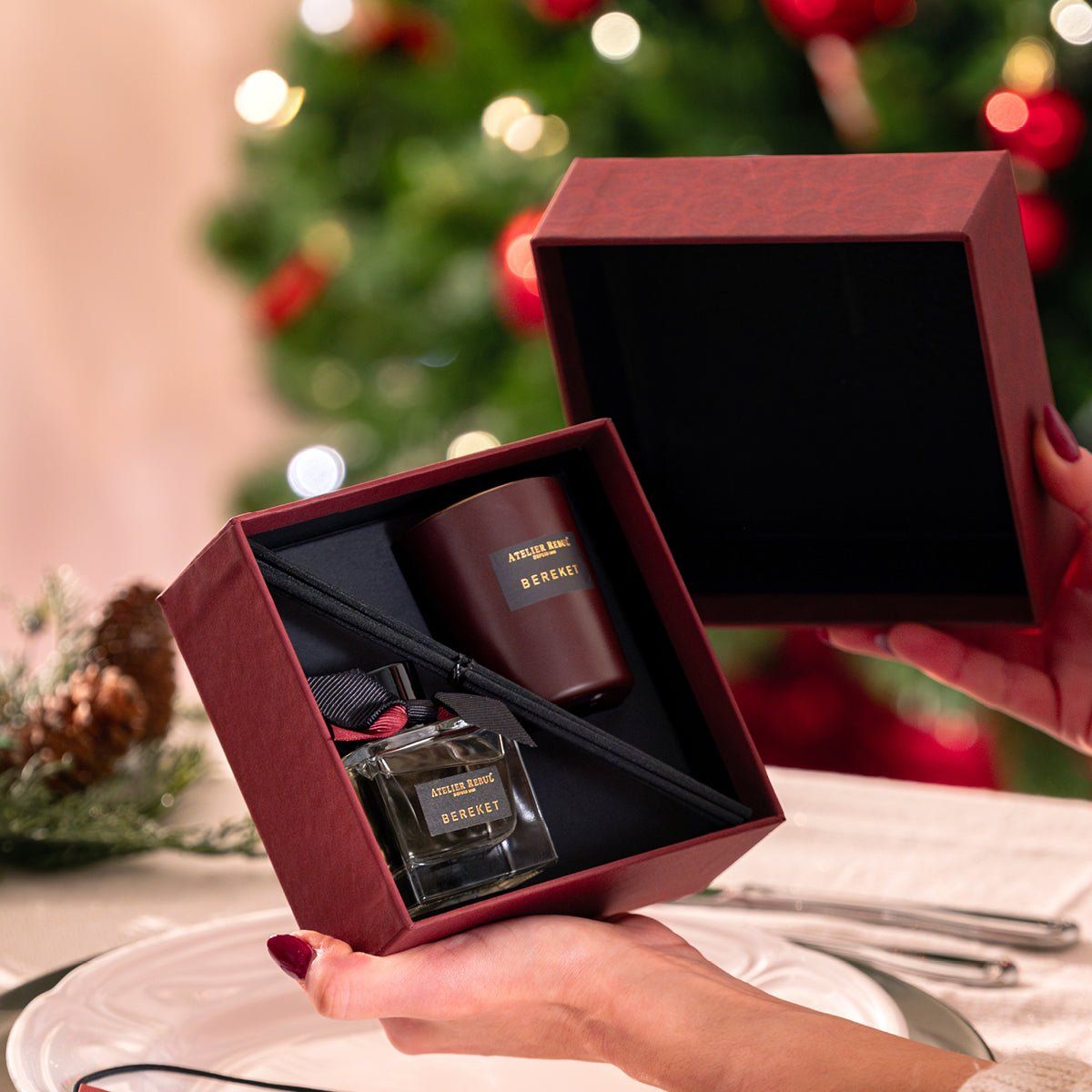 Geschenkset mit Duftdiffusor, Kerze und Flakon in offener, burgunderroter Box. Hände halten die Box vor Weihnachtsbaum.