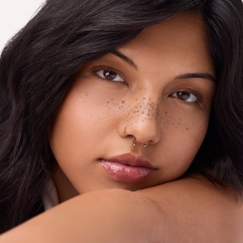 Porträt einer Frau mit Lipgloss auf den Lippen. Sie hat Sommersprossen und einen Nasenring.