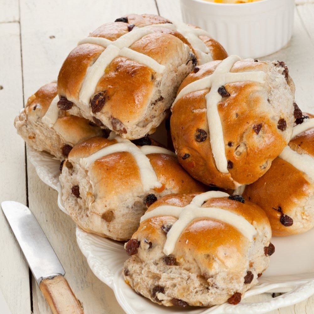 Mehrere Hot Cross Buns auf einem Teller. Die Brötchen sind goldbraun und mit einem weißen Kreuz verziert. Rosinen sind sichtbar.