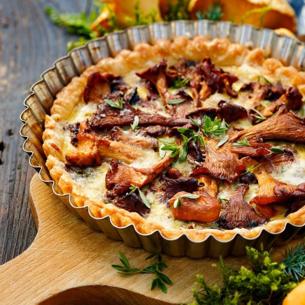 Quiche mit Pilzen in einer runden, gezahnten Form. Auf einem Holzbrett. Dekoration mit Kräutern.