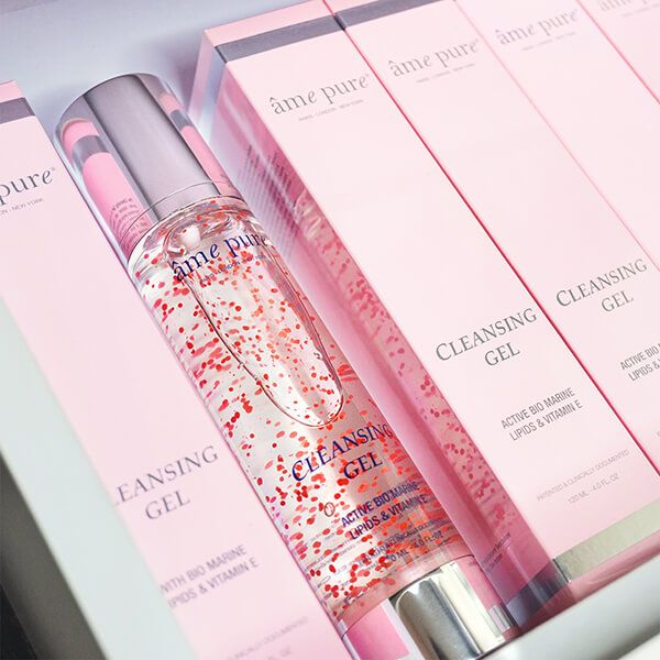 Meerdere roze verpakkingen en een fles met "âme pure" en "Cleansing GEL" tekst. Rode deeltjes zichtbaar.