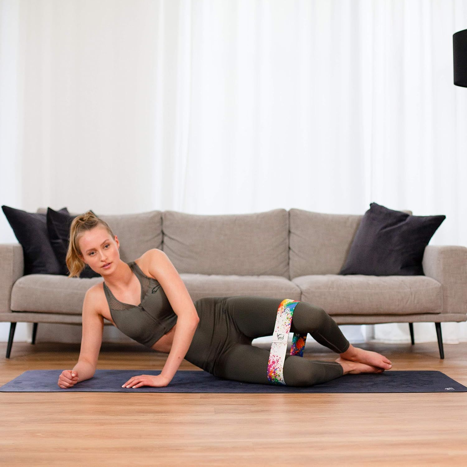 Frau liegt auf Matte, Hüftband um die Oberschenkel. Sportkleidung. Hintergrund: Sofa, Vorhang. Band mit Farbspritzern.
