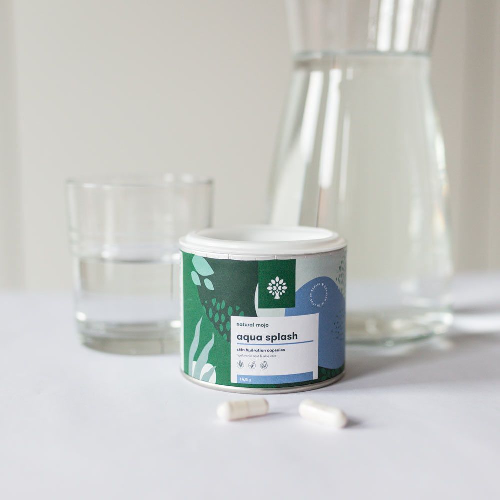 Dose "aqua splash" neben Glas, Kanne und Kapseln. Weiß-grüne Dose mit Logo. Zwei Kapseln liegen daneben.