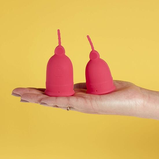 Zwei pinkfarbene Menstruationstassen auf gelbem Hintergrund. Eine Hand hält die Tassen.
