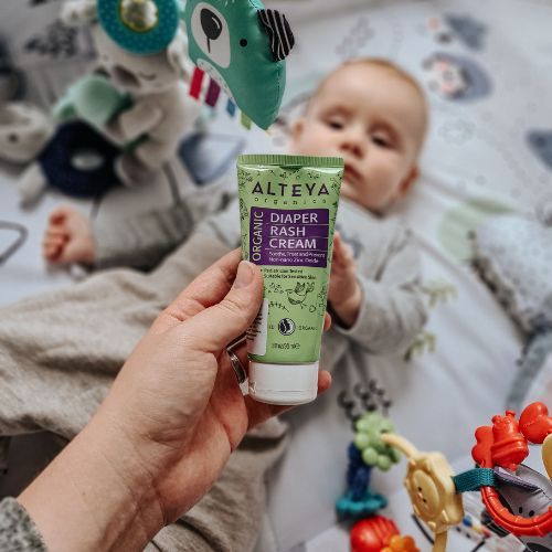 Hand hält grüne Tube 'Diaper Rash Cream'. Baby im Hintergrund, Spielzeug. Bio-Zertifizierung sichtbar.