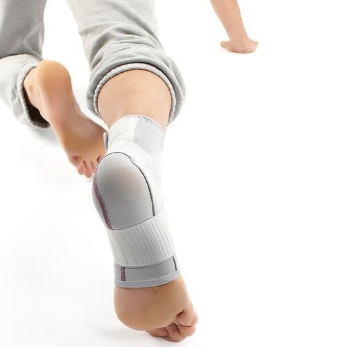 Knöchelbandage, grau-weiß. Bandage am Fuß getragen. Klettverschlüsse und Verstärkungen sichtbar. Produkt am Fuß einer Person.