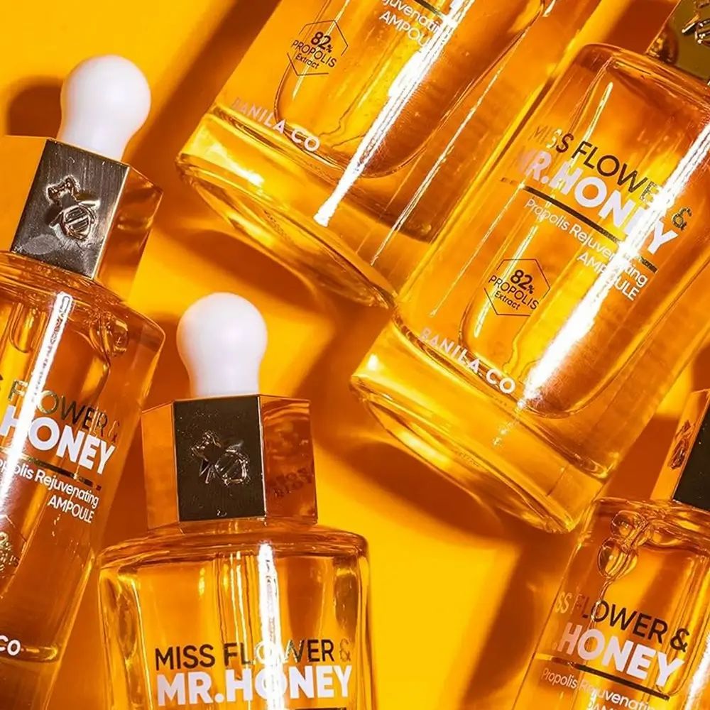 Mehrere Flaschen Gesichtsserum mit goldfarbenen Deckeln. Aufschrift: MISS FLOWER & MR. HONEY. Gelber Hintergrund.