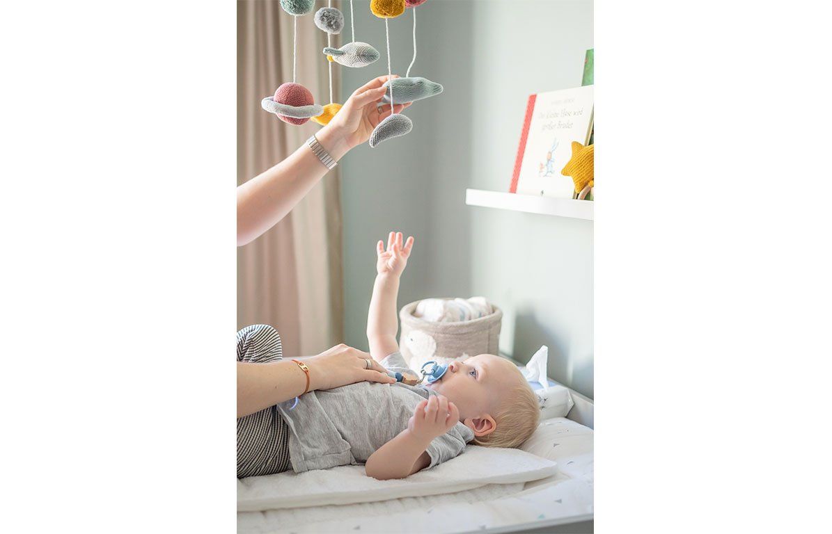 Baby liegt auf Wickelauflage, Hand greift nach Mobile. Mobile mit Holzring, bunten Kugeln, Sternen und Planeten.