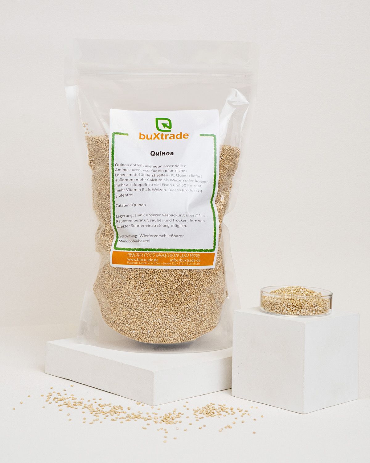 Transparente Tüte mit buXtrade Quinoa. Daneben eine kleine Schale mit Quinoa-Körnern auf einem weißen Podest. Auf dem Podest verstreute Körner.