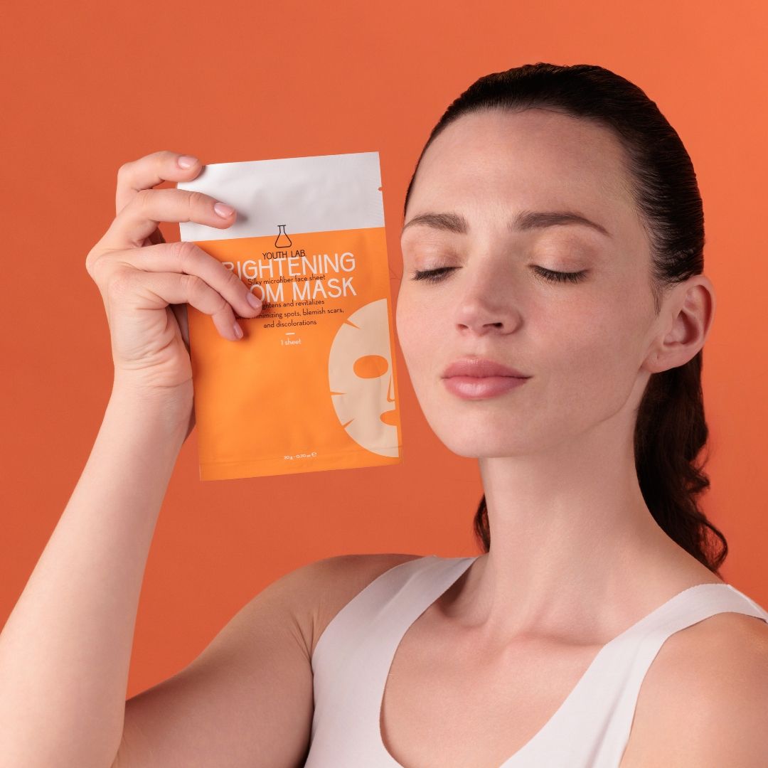 Frau hält Gesichtsmaske. Orangefarbener Hintergrund. Maske in orangefarbener Verpackung. Marke: YOUTH LAB.