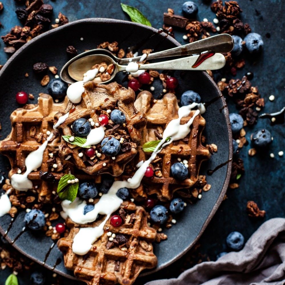 Teller mit Waffeln, Blaubeeren, Granola und Sahne. Zwei Löffel liegen auf dem Teller. Dunkler Hintergrund.