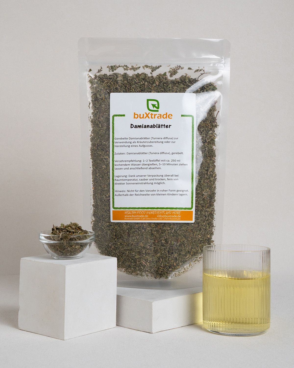 Glas mit Flüssigkeit, Kräuter in Schale und Verpackung mit Kräutern. Auf weißem Podest.