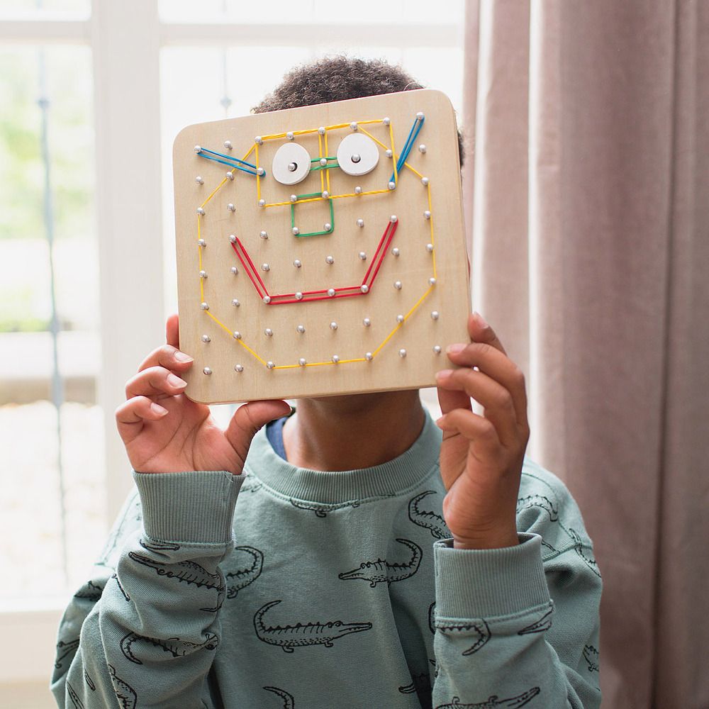 Kind hält Geoboard vor Gesicht. Holzbrett mit Gummibändern zu Gesicht geformt.