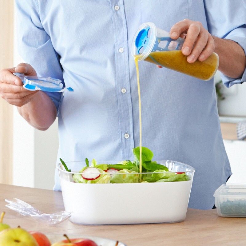 Person gießt Flüssigkeit aus einem Shaker in eine Schüssel mit Salat. Ein weiterer Shaker-Deckel liegt daneben. Auf dem Tisch liegen Äpfel.