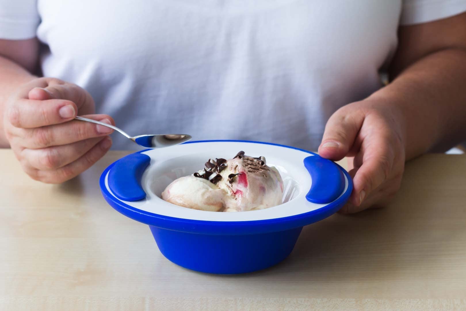 Blaue Thermoschale mit Eiscreme. Zwei Kugeln Eis, Schokoladenstückchen. Person hält Löffel. Blaue Griffe.