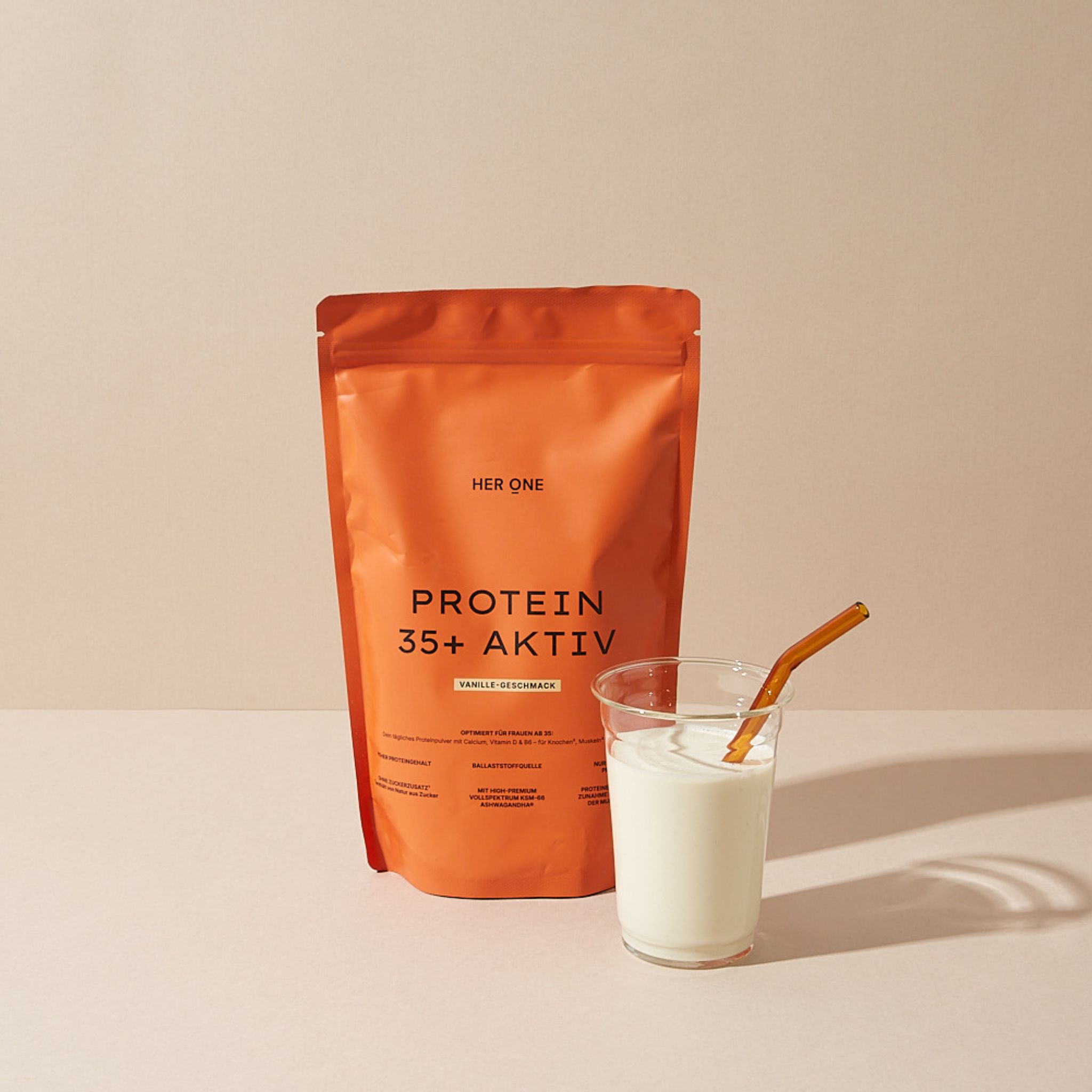 Orangefarbene Verpackung mit HER ONE, PROTEIN 35+ AKTIV. Glas mit Milch und Strohhalm daneben.