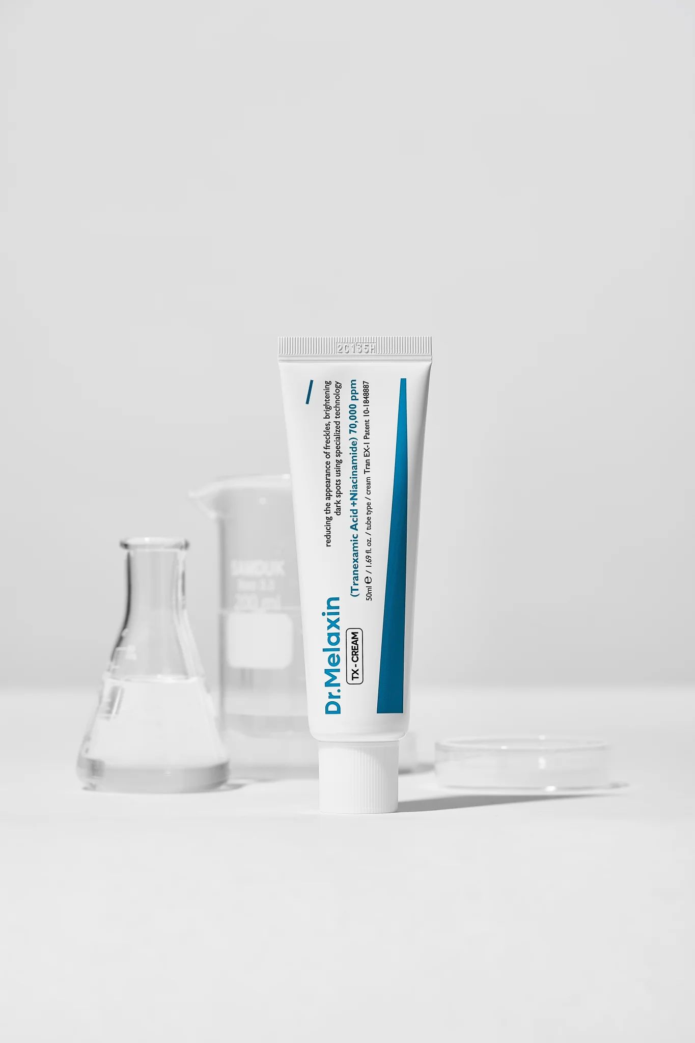 Weiße Tube neben Laborgeräten. Aufschrift: Dr.Melaxin TX-CREAM. Enthält Tranexamsäure und Niacinamid.