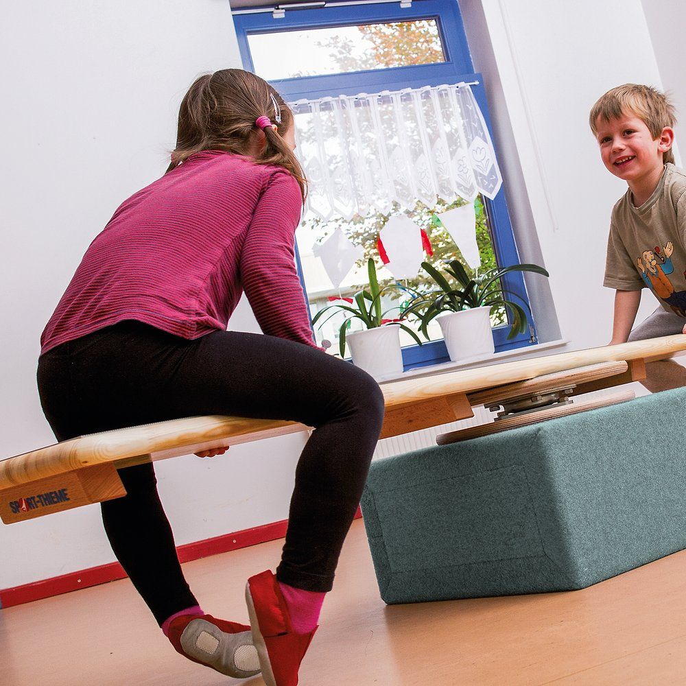 Kind balanciert auf Holzbrett mit Sport-Thieme-Logo. Ein weiteres Kind steht daneben. Im Hintergrund ein Fenster mit Vorhang.