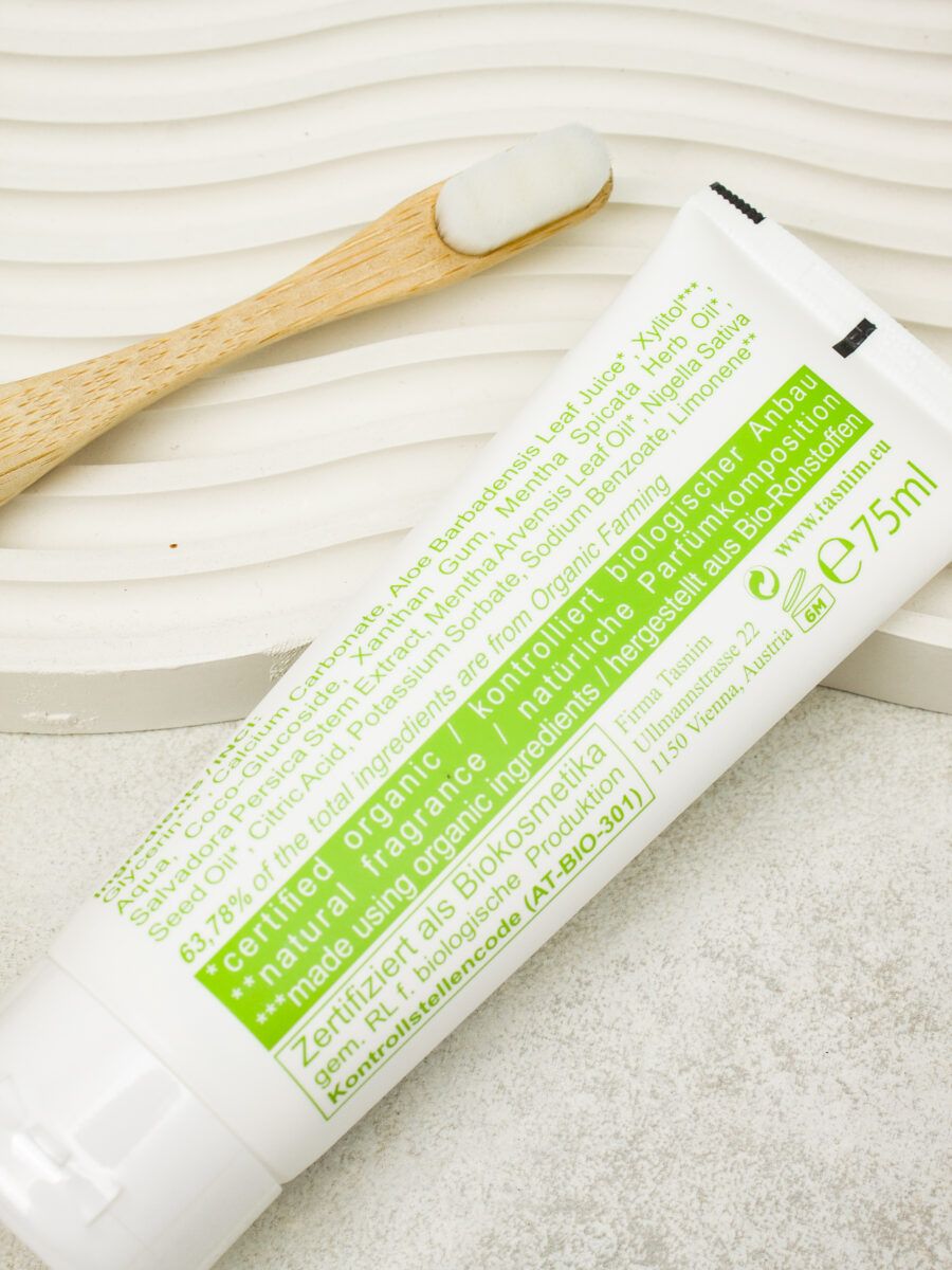 Nahaufnahme einer weißen Zahnpasta-Tube mit Text und grüner Grafik. Bambus-Zahnbürste mit Zahnpasta daneben.