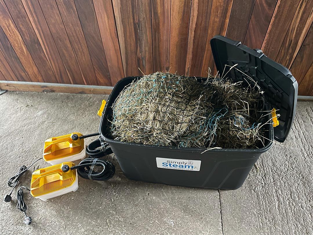 Schwarzer Behälter mit geöffnetem Deckel, gefüllt mit Heu. Simply Steam-Logo. Zwei gelb-weiße Geräte mit Kabeln. Holzverkleidung im Hintergrund.