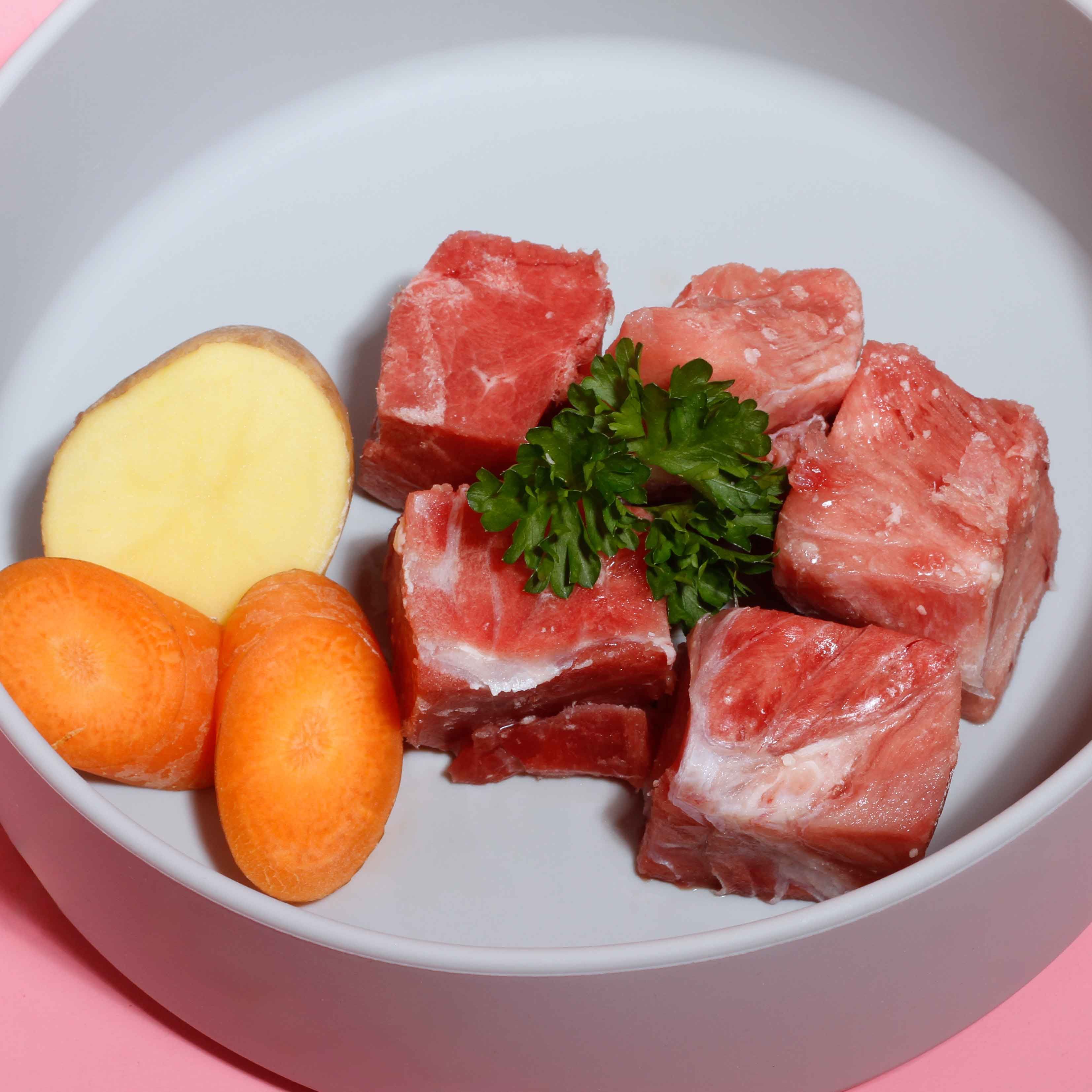 Helle Schüssel mit Fleischstücken, Kartoffelscheibe, Karotten und Petersilie.