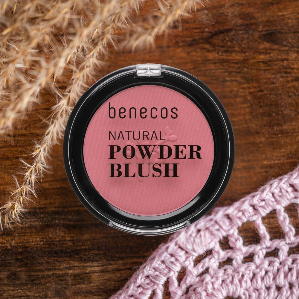 Kompakter Blush von benecos auf Holzuntergrund. Rosa Puder in schwarzem Behälter. Schriftzug: NATURAL POWDER BLUSH. Dekoration.