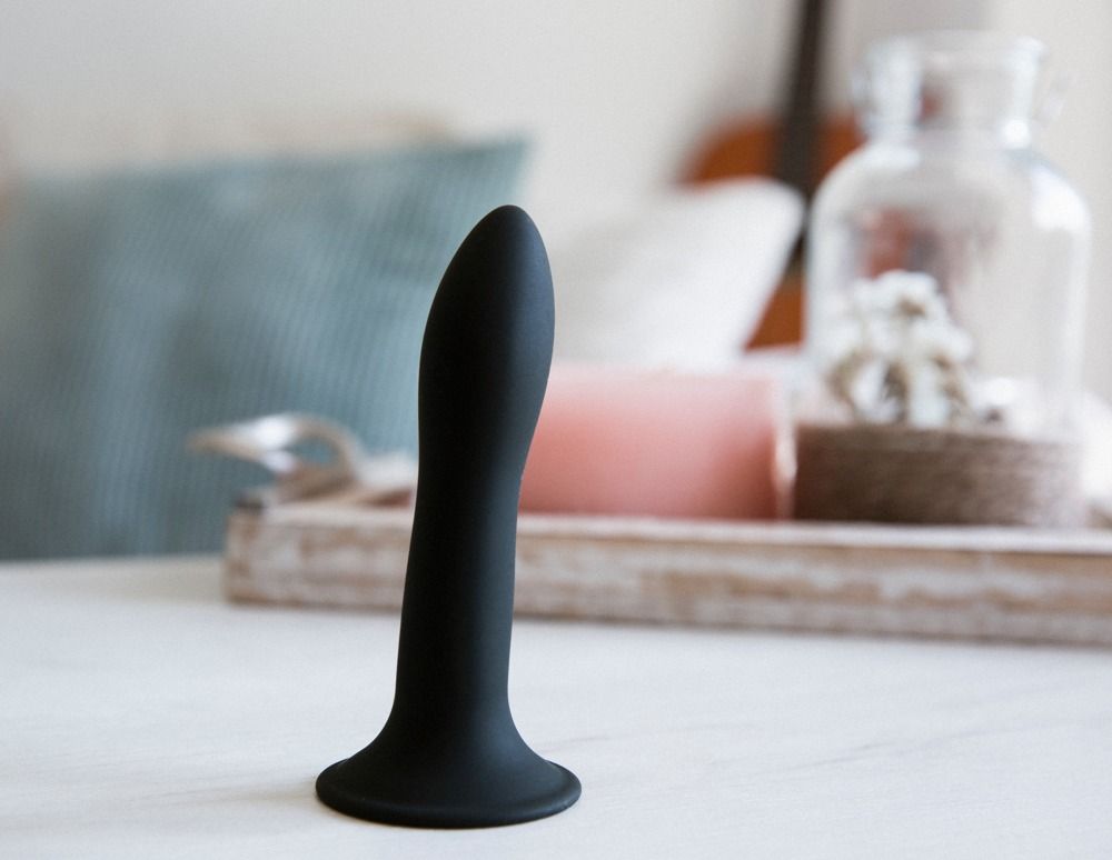 Schwarzer Dildo auf weißer Oberfläche. Breiter Fuß, geschwungene Form. Im Hintergrund: Dekoration.