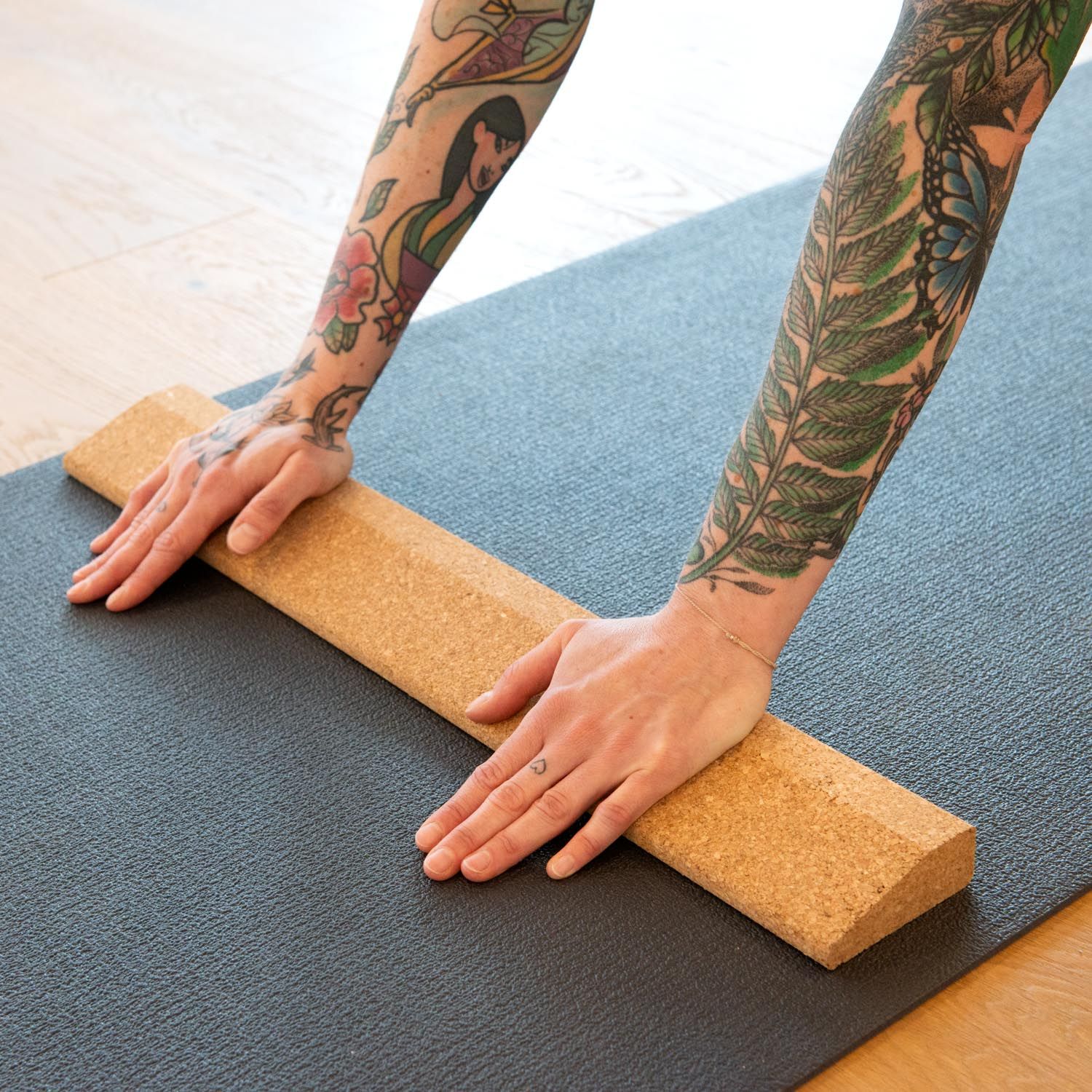 Person hält die Kork-Handstandplanke auf einer Yogamatte. Arme mit Tätowierungen. Braune Farbe.