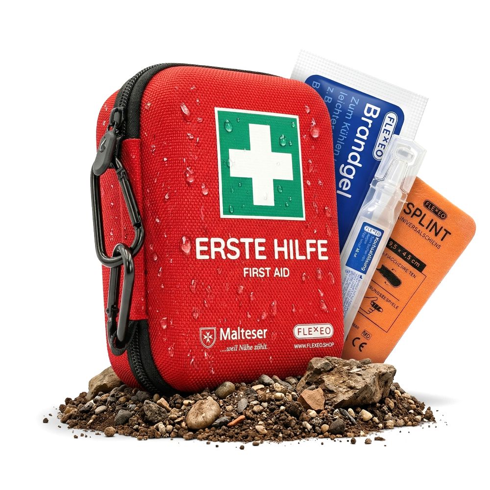 Rotes Erste-Hilfe-Set mit weißem Kreuz, schwarzem Reißverschluss und Karabinerhaken. Enthält Verbandsmaterial, Brandgel und Schiene. Auf Steinen platziert.