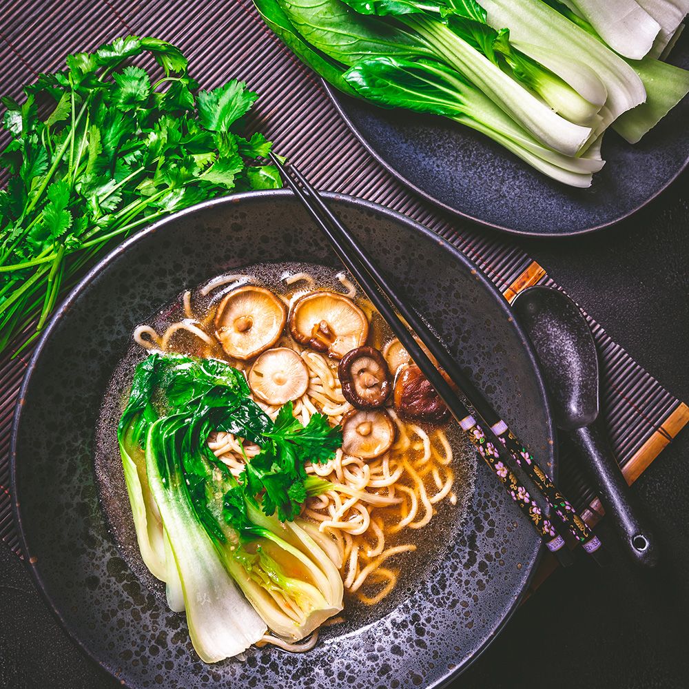 Schwarze Schüssel mit Nudelsuppe, Pilzen, Bok Choy und Koriander. Essstäbchen und Löffel liegen daneben. Auf Bambusmatte.